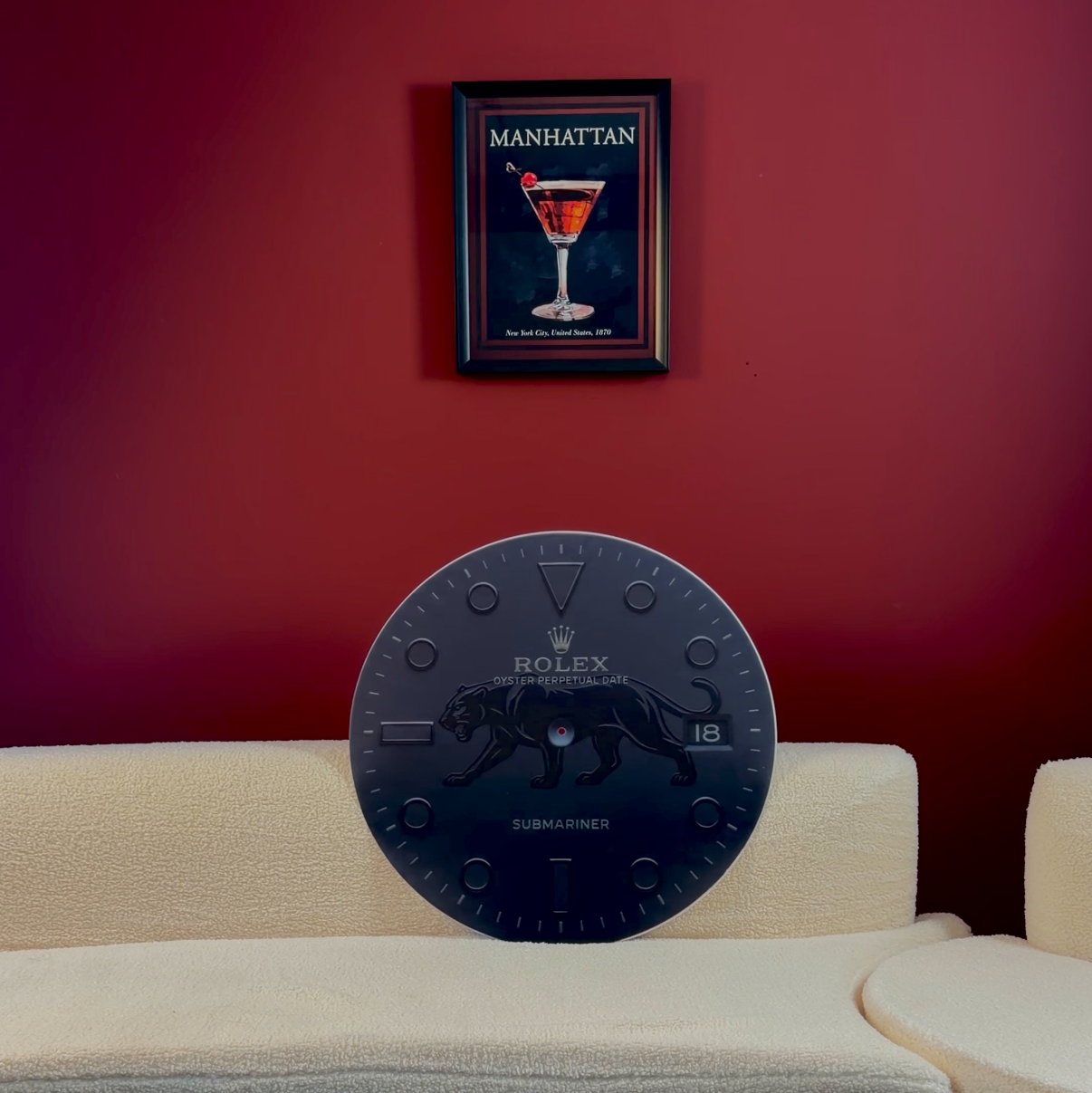 A dark round clock with Roman numerals displayed on a cream-colored sectional sofa in front of a deep red wall, with a framed 'The Art of Martini' print hanging above.